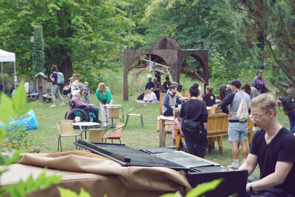 Gesellige Stimmung im Garten bei Krakeele. Im Vordergrund spielt ein Gast auf dem Flügel auf der Terrasse.