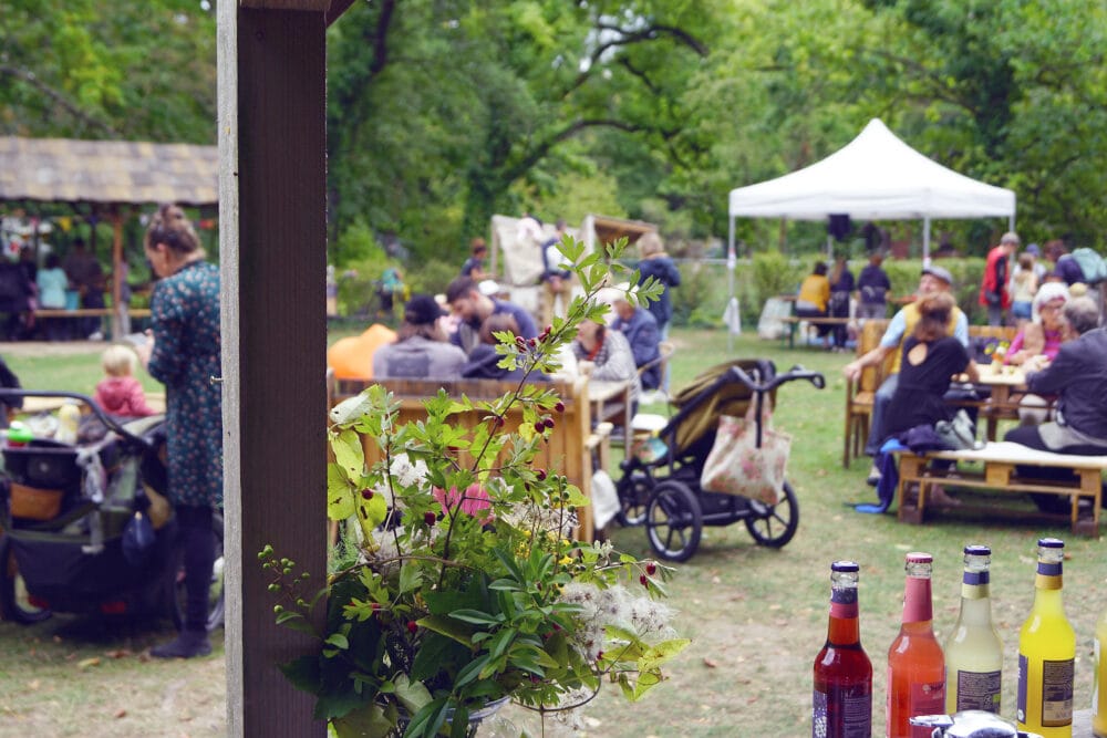 Im Garten vom Haus 4 sehen wir das gesellige Publikum von Krakeele. Im Vordergrund stehen Getränke und ein schöner Blumenstrauß auf einer Theke.