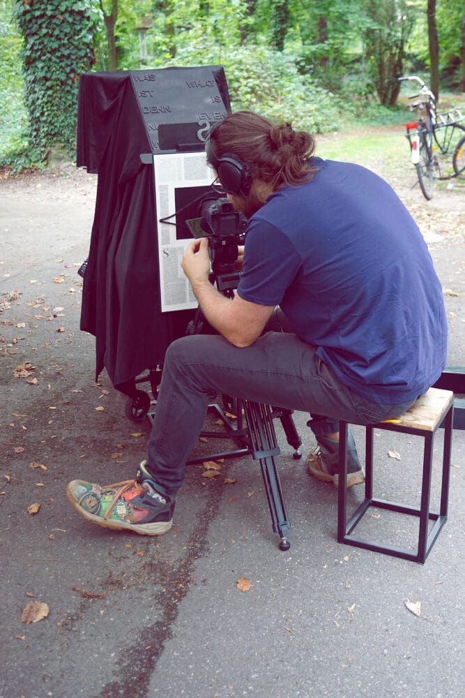 Ein Besucher filmt mit seiner Kamera die Vorstellung beim Minitheater durch ein Schauloch, in einem Kasten. Auf der anderen Seite spielt eine Puppenspielerin für die Zuschauer ein kleines Stück.