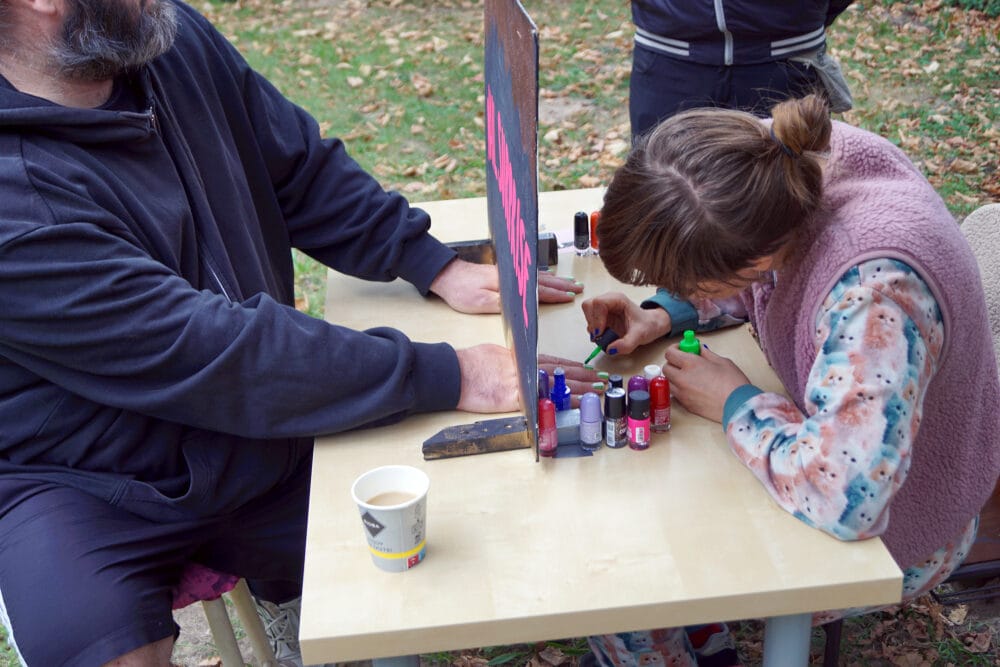 Ein Mann lässt sich bei Nail Surprise die Nägel verzieren. Dabei muss man sich vom Ergebnis überraschen lassen, denn während dem Lackieren kann man nicht sehen was passiert, da die Hände durch eine Öffnung in einem Brett gesteckt werden müssen.