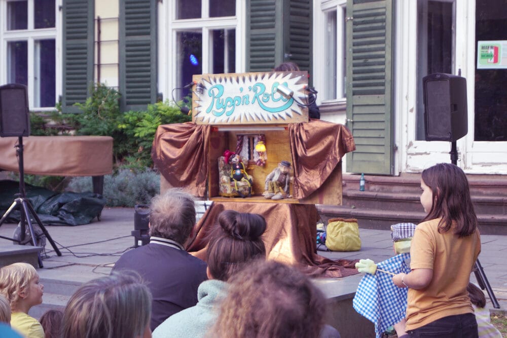 Mit dem Publikum sehen wir die witzig gestaltete Bühne von Pupp'n'Roll. 2 Puppen sitzen in einem Wohnzimmer und sehen sich ihre Lieblingsshow im Fernsehen an.