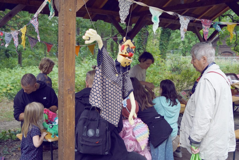 Eine Seniorin betrachtet das bunte Treiben an einem Stand, an dem Kinder und Erwachsene Puppen zum Puppenspiel bauen können.