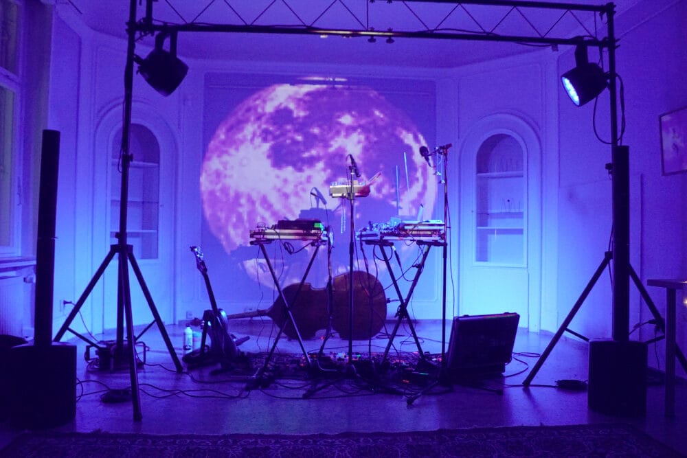 Es ist eine Konzertbühne zu sehen. Die Bühne ist vor Beginn des Konzerts noch leer und ganz blau ausgeleuchtet. Im Hintergrund der Bühne wurde ein großer Mond an die Wand projiziert.