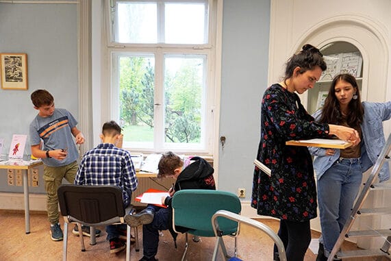 Jugendliche bereiten eine Ausstellung vor. Rechts im Bild rahmen zwei personen ein Bild, um es aufzuhängen, Links im Bild schreiben Jugendliche Beschreibungstexte für die Bilder und eine Eröffnungsrede für die Ausstellungseröffnung.