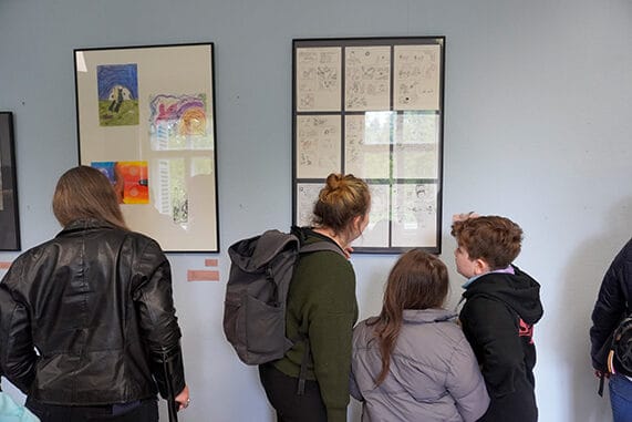 Besucher*innen stehen vor den Bildern der Ausstellung "Galerie der 2000 Ideen" und betrachten Zeichnungen in zwei Rahmen.