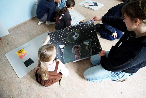 Zwei Personen betrachten ein gemaltes Bild, im Hintergrund sind Kinder zu sehen, die sich die Bilder in einer Mappe ansehen.