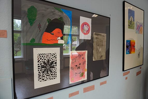 Auf dem Bild sind zwei größere Bilderrahmen zu sehen, in denen mehrere kleinere Formate hängen: ein Linolschnitt einer Blume, ein gemalter Fuchs in einer Höhle, eine Monotypie von einem Herz und zwei abstrakte Bilder.