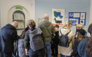 Zu sehen sind mehrere Personen, die die Ausstellungseröffnung der "Galerie der 2000 Ideen" besuchen und sich die Bilder an den Wänden und in der Vitrine ansehen.