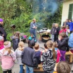 Kindergruppe steht um ein Feuer auf einer Terrasse. Über dem Feuer ist ein Schwenkkessel mit Essen.