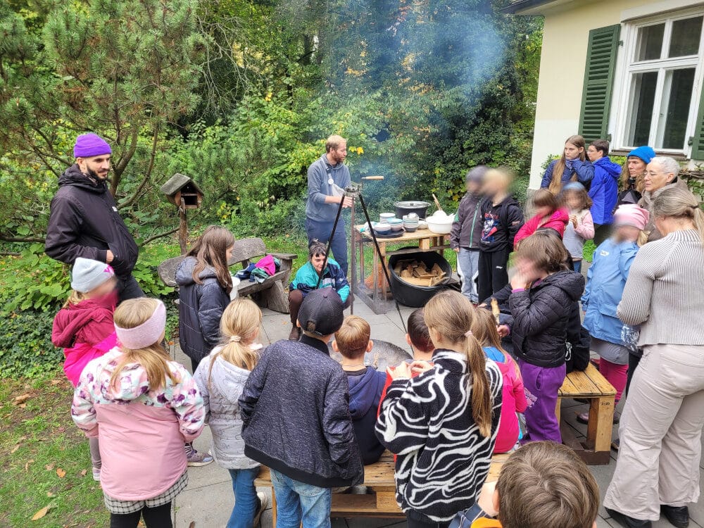 Kindergruppe steht um ein Feuer auf einer Terrasse. Über dem Feuer ist ein Schwenkkessel mit Essen.