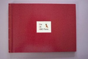 Dunkelrotes Buchcover mit Reptilien-Struktur und orangener Bindung, das den Buchtitel und eine kleine Kinderzeichnung eines orangenen Tieres zeigt