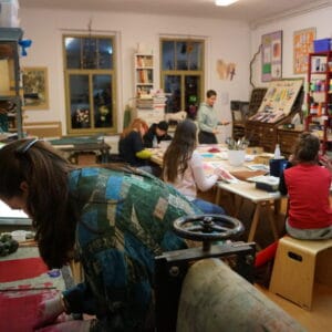 Im Atelier sitzen mehrere Kursteilnehmer*innen an einem großen Tisch und zeichnen, zwei Personen unterhalten sich über Bilder und im Vordergrund arbeitet eine Person an Monotypien.