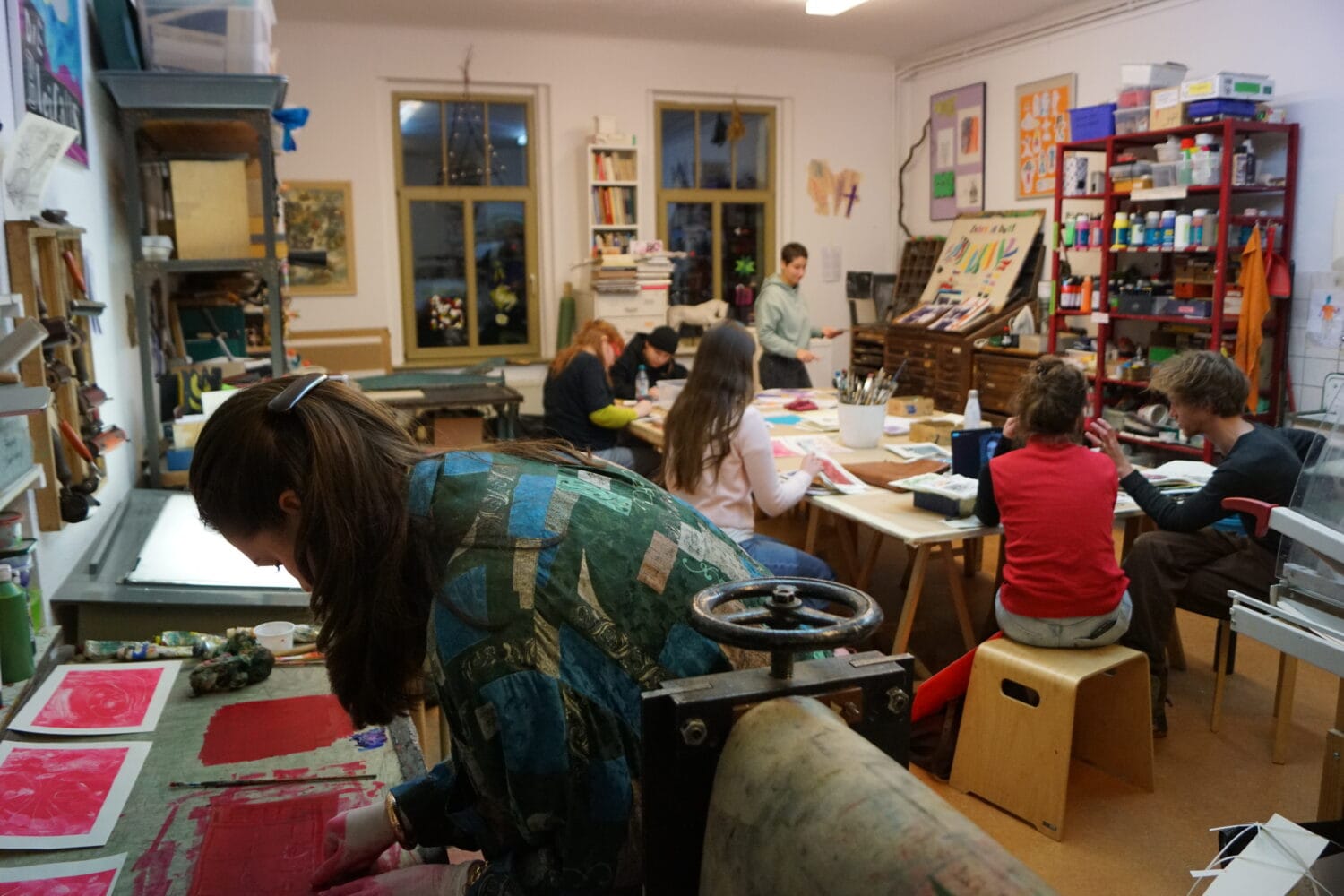 Im Atelier sitzen mehrere Kursteilnehmer*innen an einem großen Tisch und zeichnen, zwei Personen unterhalten sich über Bilder und im Vordergrund arbeitet eine Person an Monotypien.